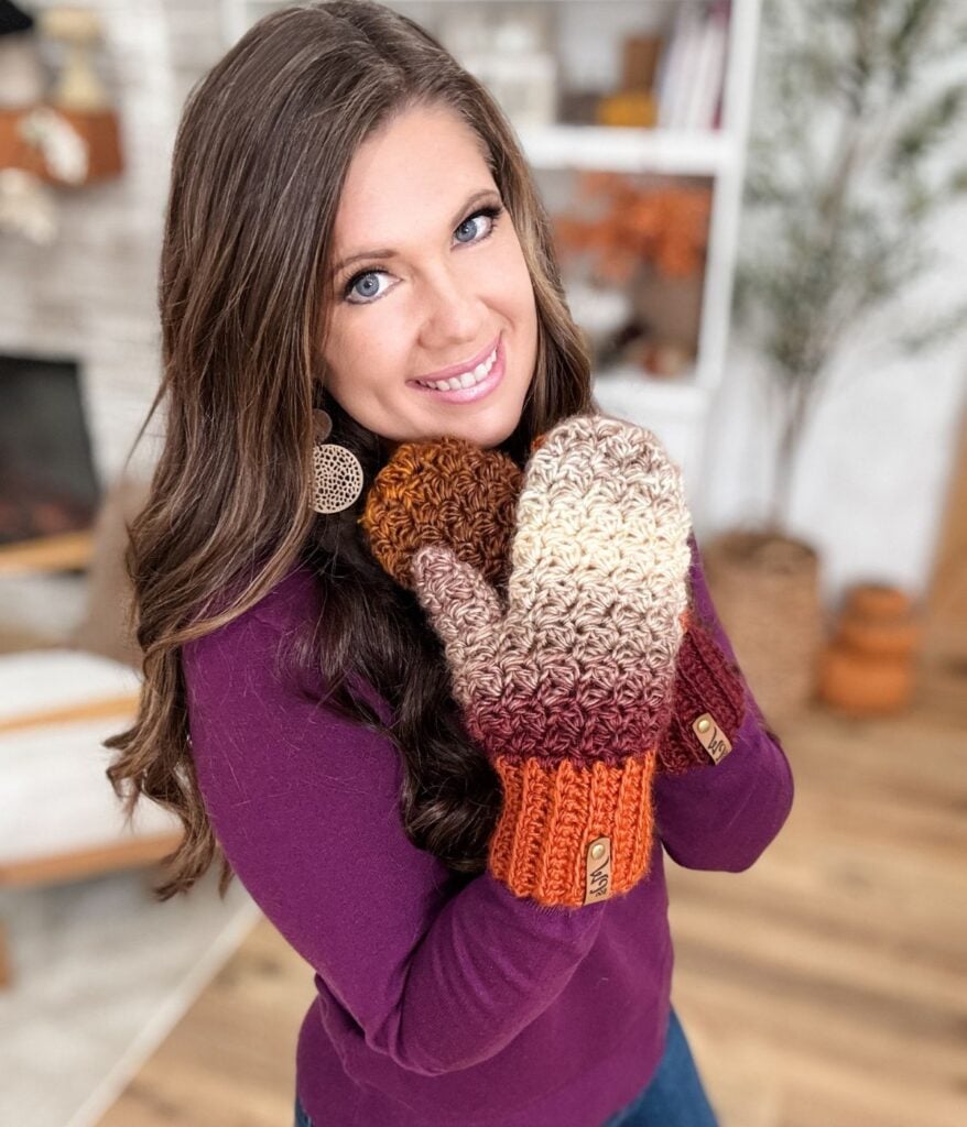 Woman with long brown hair wears a purple sweater and gradient knit fingerless gloves and mittens, smiling and posing indoors in a cozy, well-lit room.