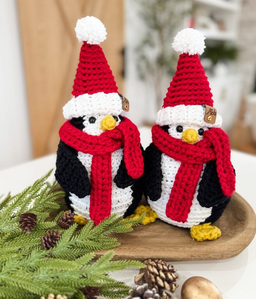 Two crocheted Santa Penguin figures wearing red hats and scarves sit on a wooden tray next to pine branches and pine cones.