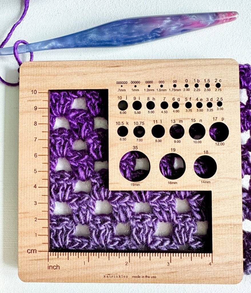 A wooden crochet gauge tool measures purple crocheted fabric, with a blue and pink crochet hook and yarn above the tool—perfect for following a beginners guide to achieving accurate crochet results.