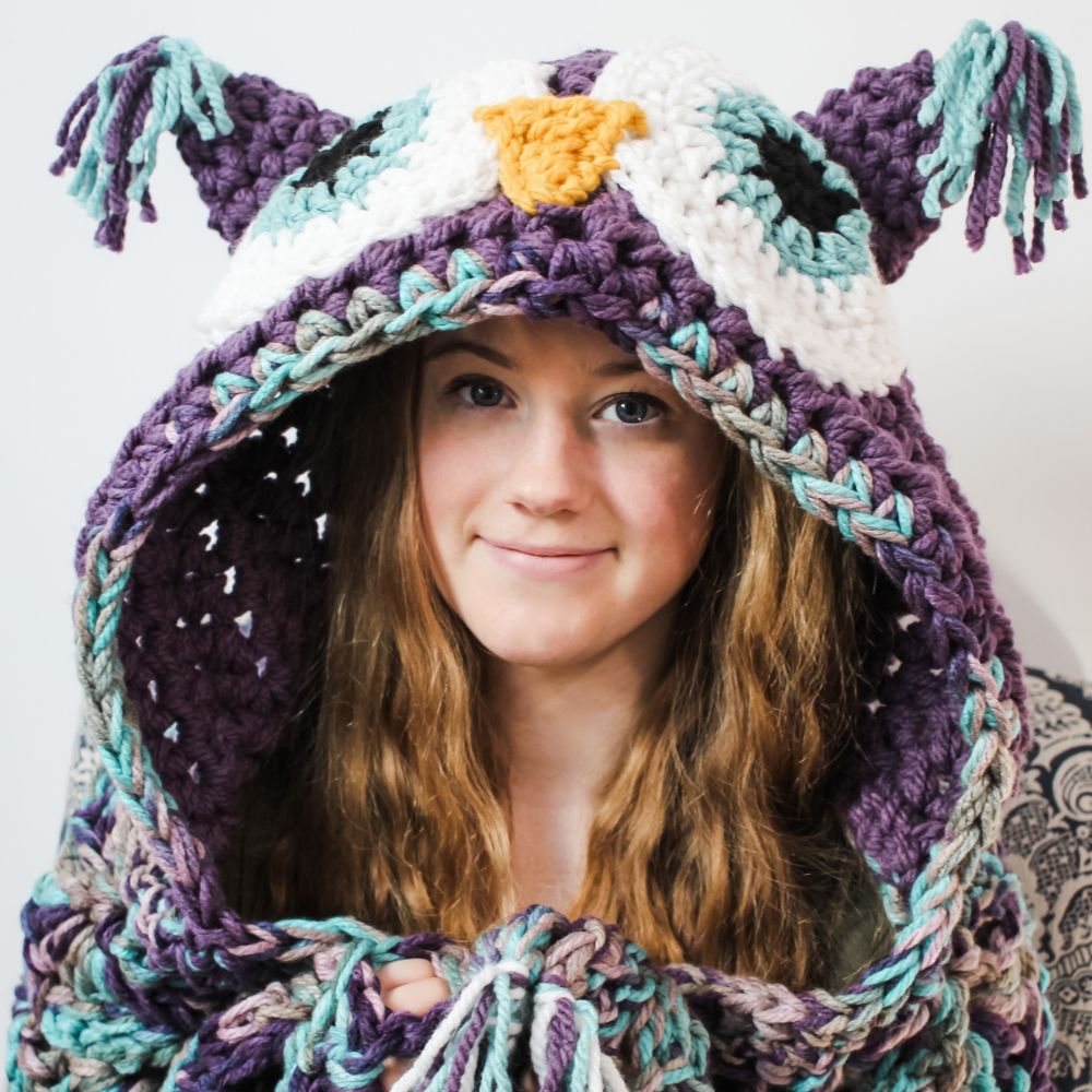 A person with long hair wears a purple, crocheted owl hooded blanket with large eyes, a beak, and tassel details.