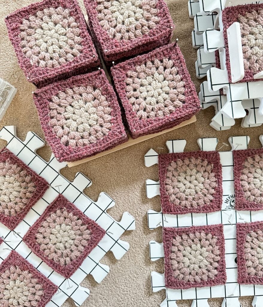 Several crocheted granny squares in pink and beige are laid out on a surface, with some stacked in a box and others pinned to blocking mats for shaping—perfect for following along with a beginners crochet guide or checking your crochet gauge.