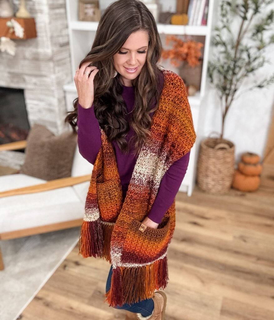 A woman with long brown hair wearing a purple top and a cozy pocket shawl with fringe stands in an autumn-themed living room.