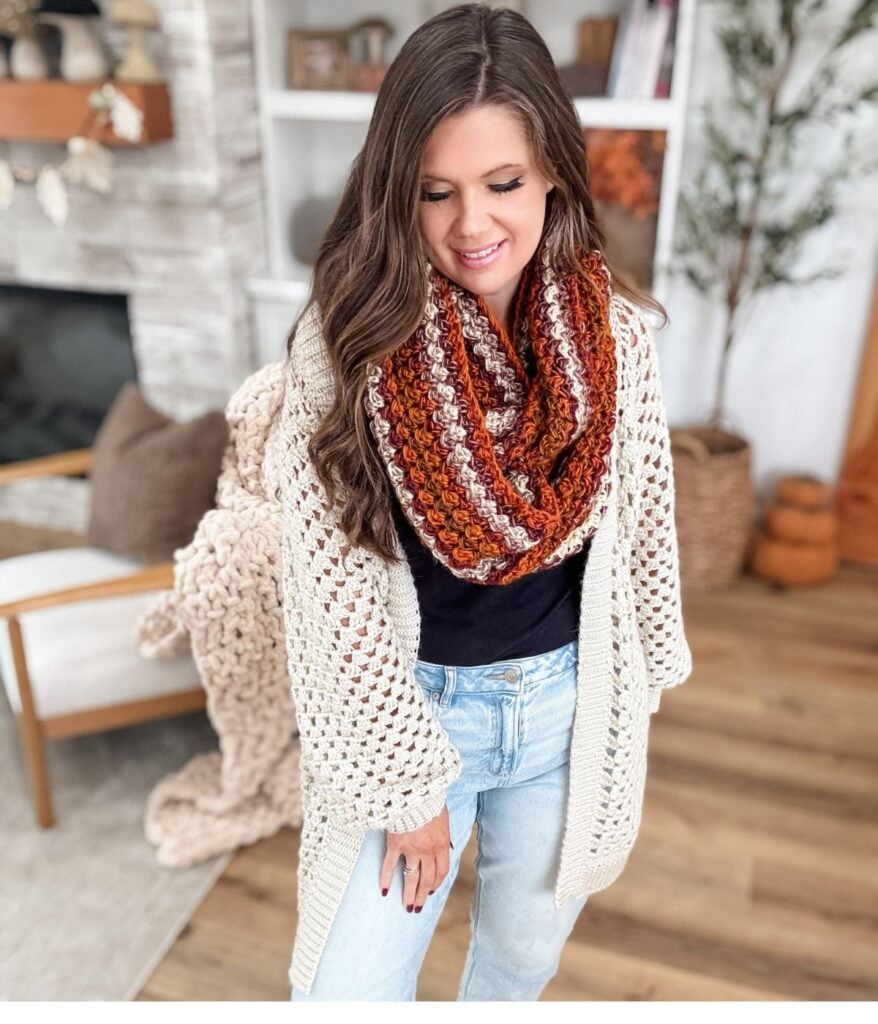 A woman wearing a chunky knit cardigan and an infinity scarf stands in a cozy, modern living room with neutral decor.