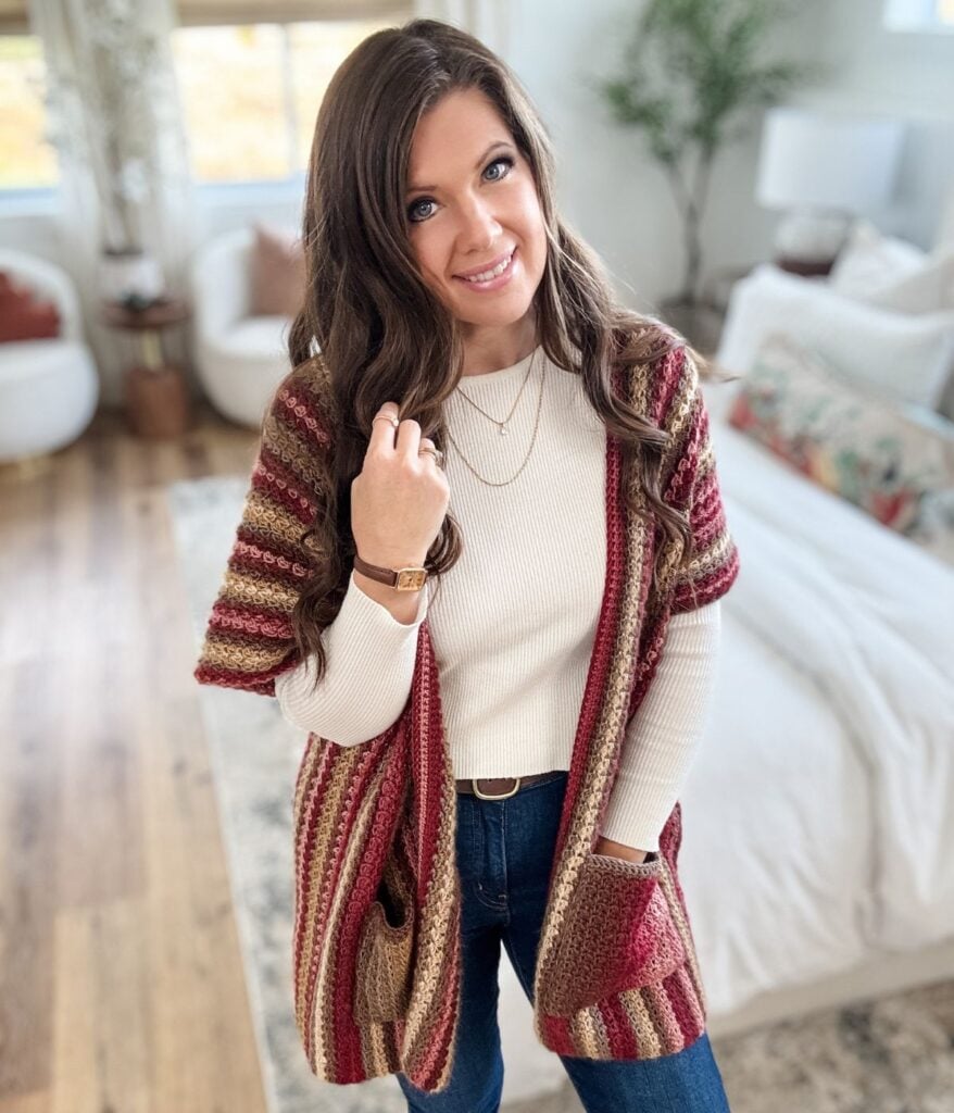 A woman with long brown hair, wearing a cream top, jeans, and a Suzette Ruana striped cardigan, stands in a bright, modern living room with wood floors and white furnishings.