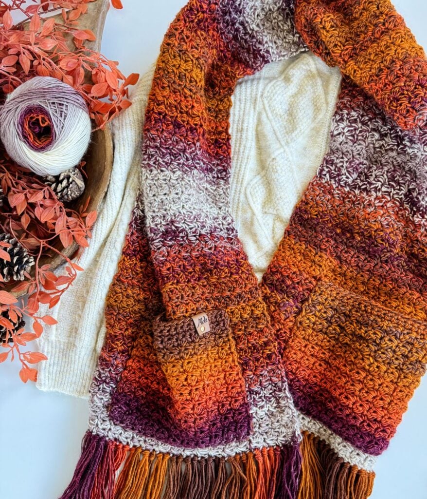 A multicolored pocket shawl with fringe, in shades of orange, purple, and cream, is laid out beside a cream sweater, pinecones, and autumn leaves.