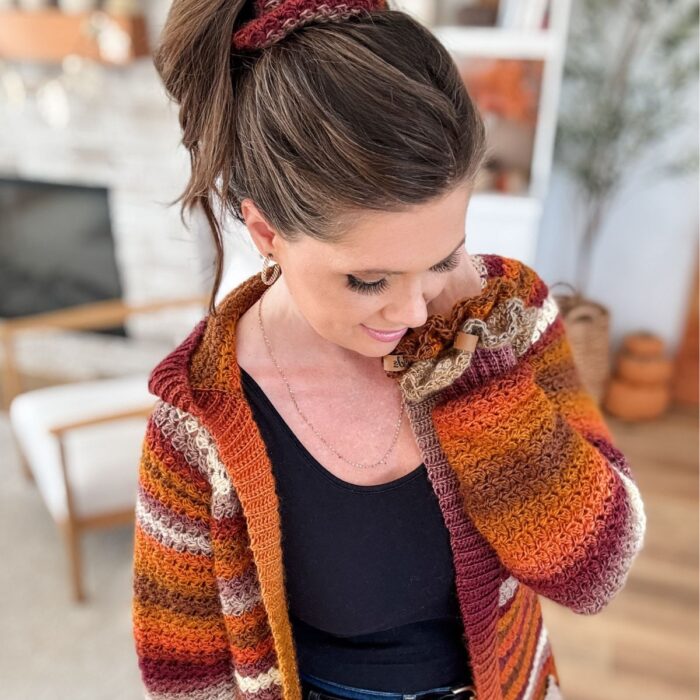 Woman wearing a striped, autumn-colored knit cardigan and a matching crochet scrunchie looks down and smiles inside a cozy, well-lit living room. Try making your own Suzette Stitch crochet scrunchie with the free pattern available!.