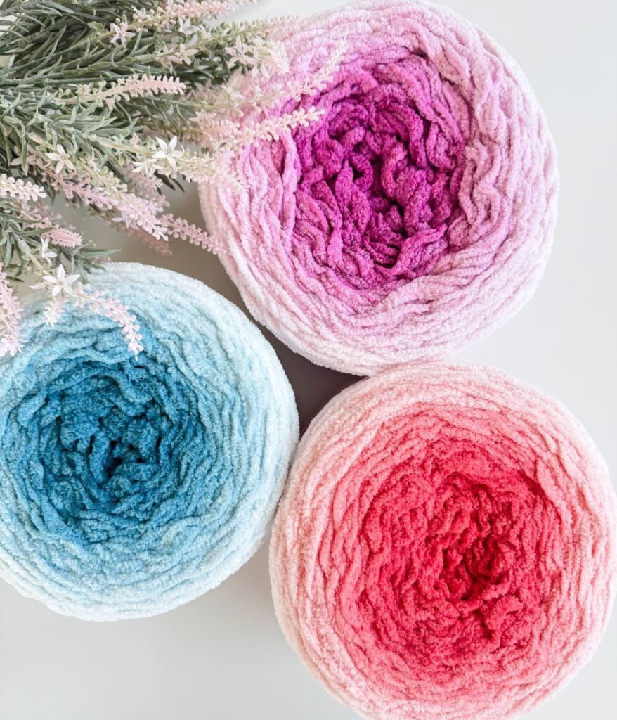 Three rolled towels in shades of pink, blue, and red are arranged in a triangle shape next to a sprig of artificial lavender flowers on a white surface—perfect for a cozy 2-day throw.