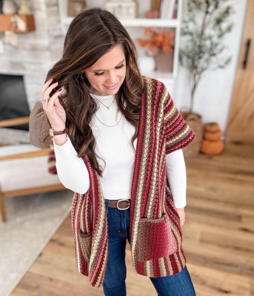 A woman wearing a striped Suzette Ruana vest with pockets over a white long-sleeve top and jeans stands in a living room with wooden floors and stylish home decor in the background.