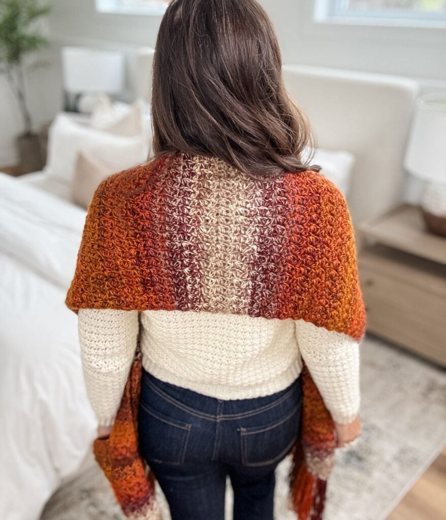 A person with long brown hair, wearing a cream sweater and dark jeans, stands indoors with a multicolored crocheted pocket shawl draped over their shoulders.