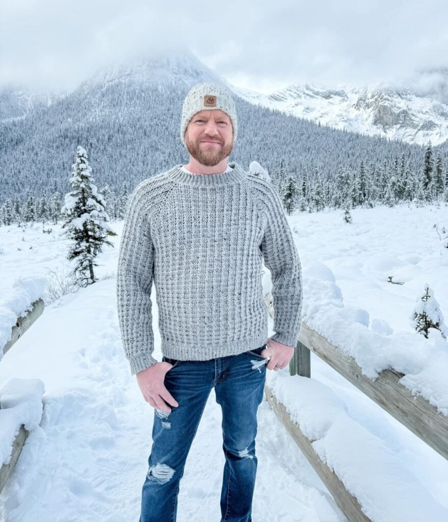A man in a gray knit sweater from MJ's Off The Hook Designs, jeans, and a beanie stands on a snowy wooden walkway with snow-covered trees and mountains in the background.