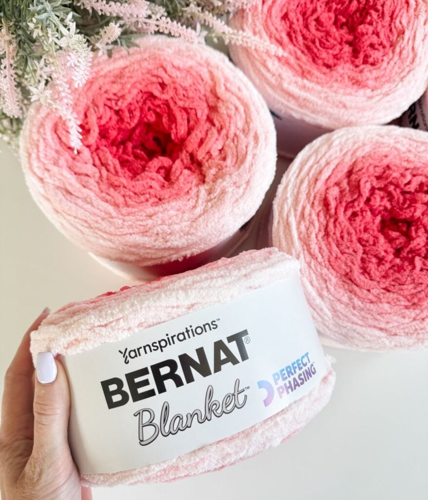 A hand holds a skein of Bernat Blanket yarn in a pink gradient, labeled "Perfect Phasing"—ideal for creating a cozy 2-day throw. Three more skeins and pink flowers are beautifully arranged in the background.