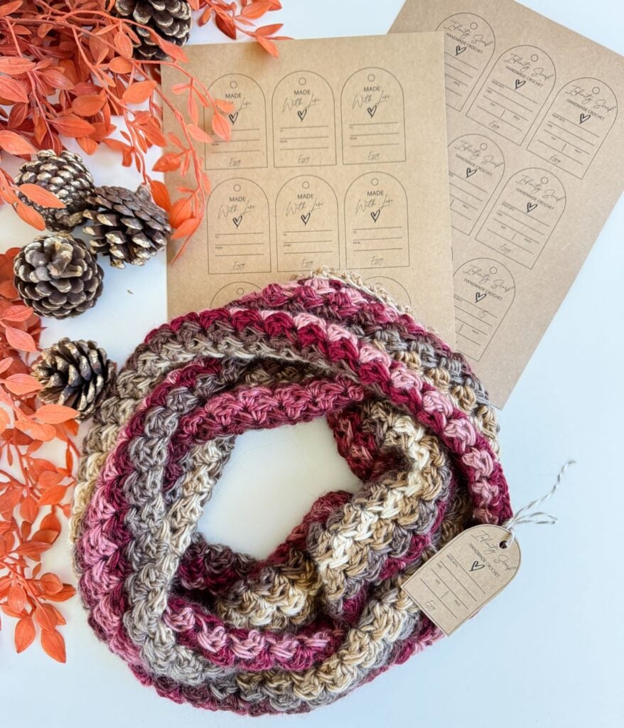 A crocheted infinity scarf in pink, beige, and brown tones with a kraft paper tag, surrounded by orange leaves, pinecones, and sheets of similar tags on a white surface.