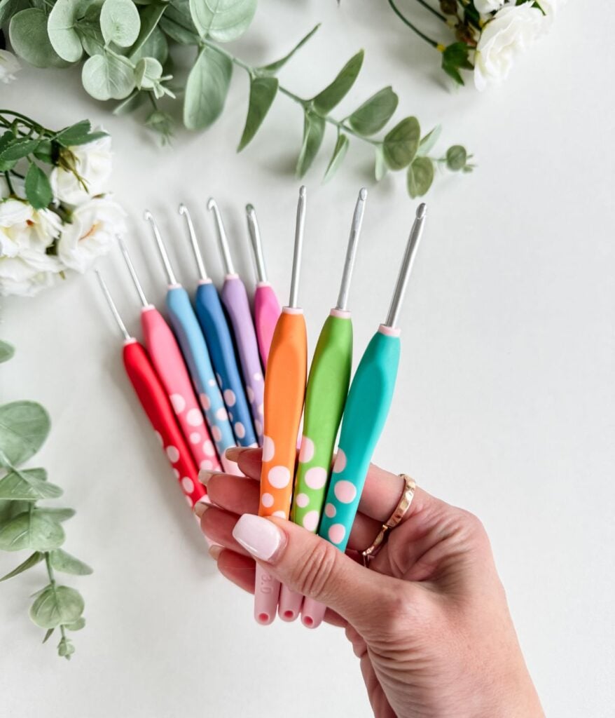 A hand holds five colorful crochet hooks with polka dot handles—perfect for any beginners guide. More crochet hooks and green foliage are visible in the background on a white surface.