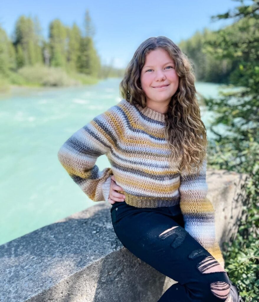 A girl with long curly hair wearing a striped sweater from MJ's Off The Hook Designs and black ripped jeans poses on a stone ledge by a river, with trees in the background.