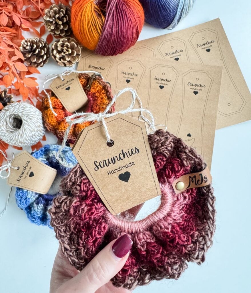 A hand holds a handmade crochet scrunchie crafted with the Suzette Stitch and a labeled tag. Other crochet scrunchies, yarn balls, pinecones, orange leaves, and printed labels are arranged on a white surface.
