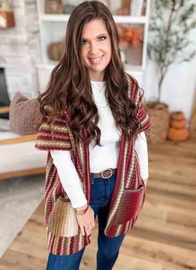 Woman with long brown hair wearing a striped Suzette Ruana knit cardigan, white top, and jeans stands indoors on a wooden floor, smiling at the camera.