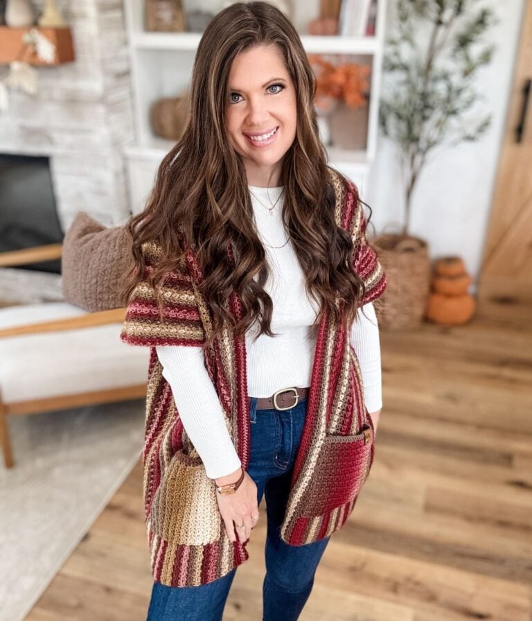 Woman with long brown hair wearing a striped Suzette Ruana knit cardigan, white top, and jeans stands indoors on a wooden floor, smiling at the camera.