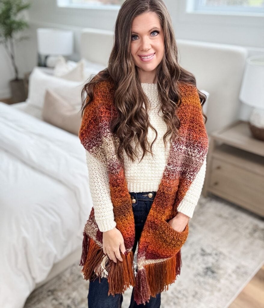 A woman with long brown hair wearing a cream sweater, jeans, and a large orange and brown fringed pocket shawl stands in a bedroom.