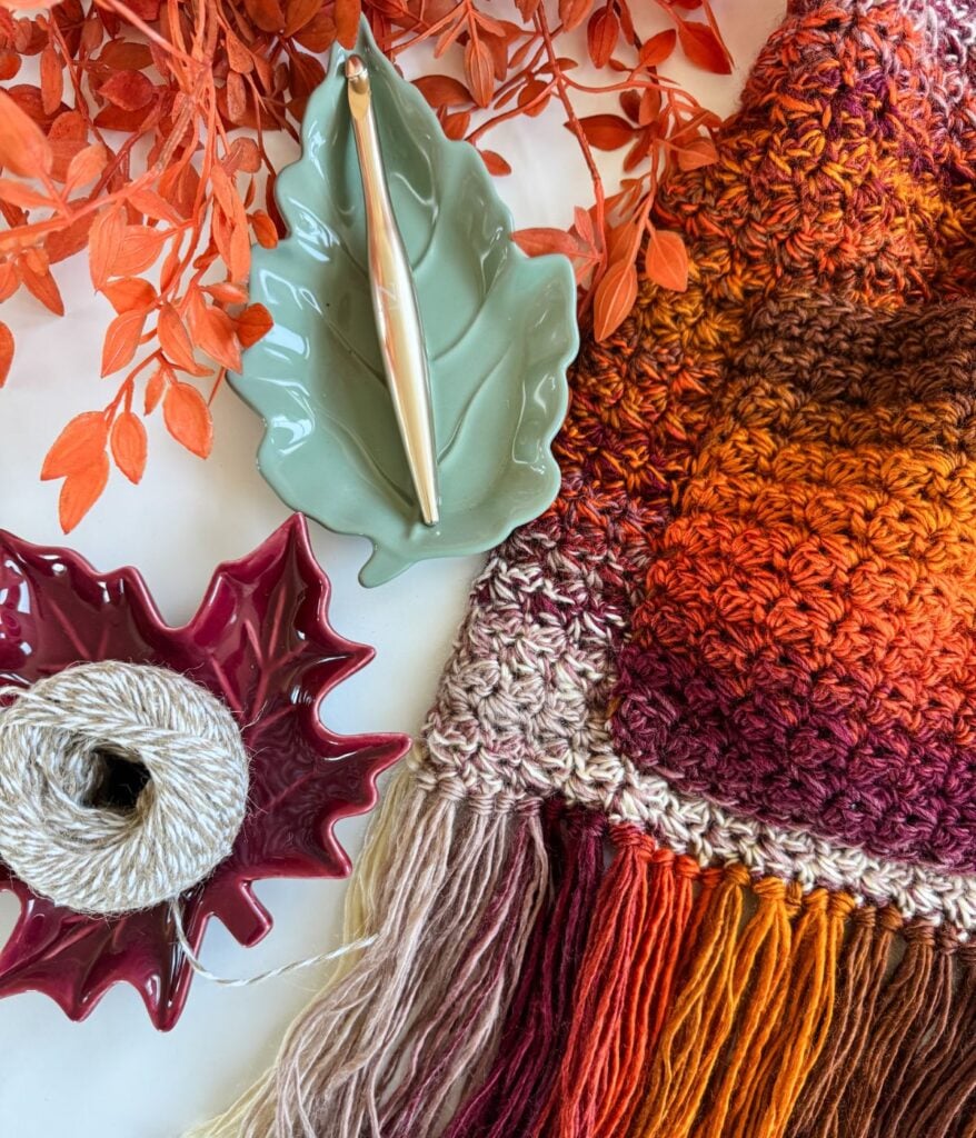 Yarn, a crochet hook on a green leaf-shaped dish, a ball of string on a red dish, orange leaves, and a multicolored crocheted pocket shawl with fringe are arranged on a white surface.