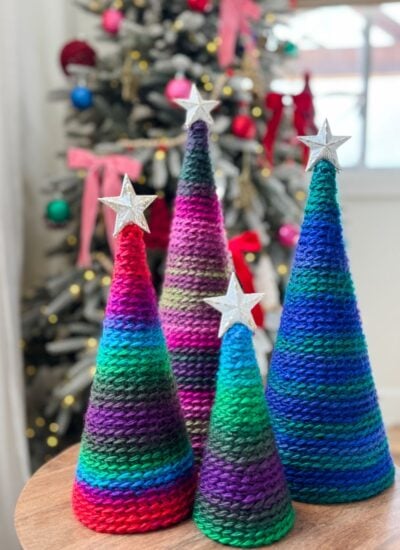 Four colorful knitted cone-shaped trees with silver star toppers are displayed on a table beside a cozy pocket shawl, with a decorated Christmas tree in the background.