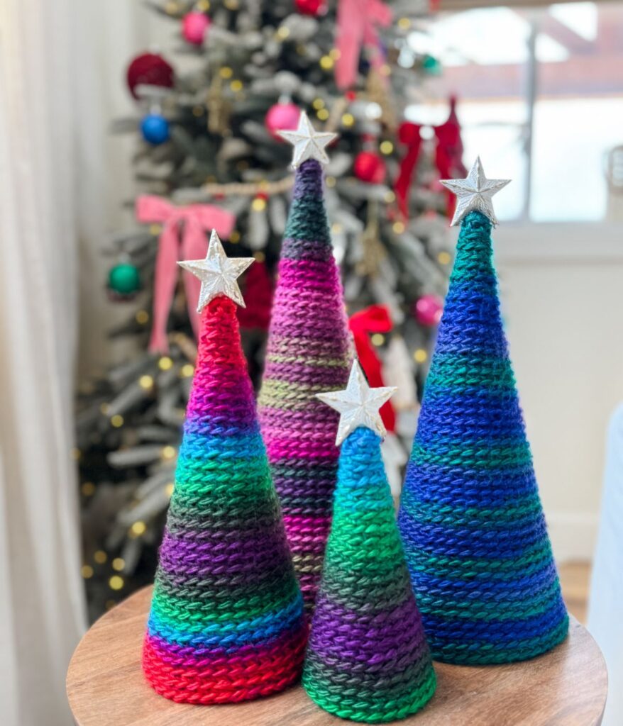 Four colorful knitted cone-shaped trees with silver star toppers are displayed on a table beside a cozy pocket shawl, with a decorated Christmas tree in the background.