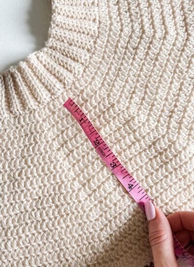 A pink measuring tape is held across the chest of a cream-colored knitted sweater, indicating a measurement—helpful for checking crochet gauge or as a beginners guide to accurate sizing.