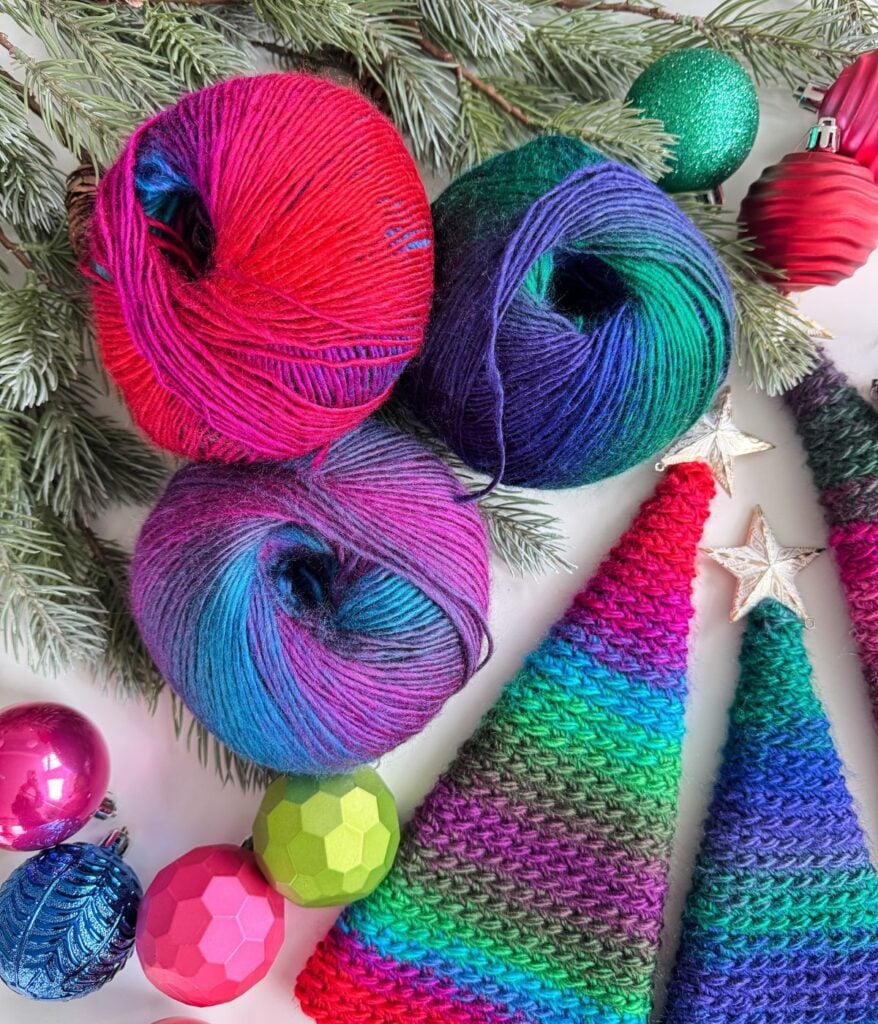 Three colorful balls of yarn, a cozy Pocket Shawl, knitted cone trees, and assorted Christmas ornaments are arranged on pine branches.