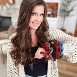 Woman with long brown hair, wearing a cream crochet cardigan and black top, displays several colorful Crochet Scrunchie creations on her wrist in a cozy living room.