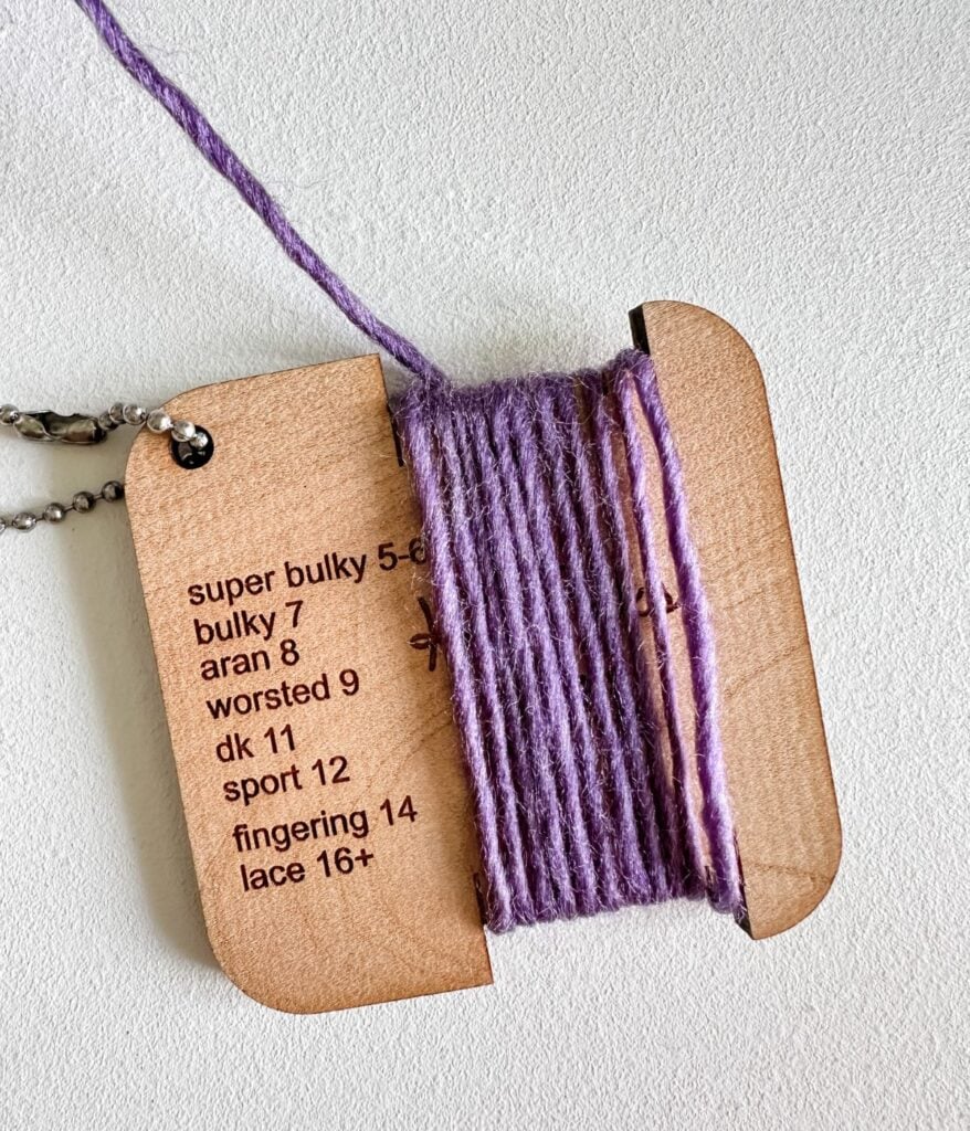 A wooden yarn gauge tool with purple yarn wrapped around, showing thickness measurements for categories like bulky, worsted, and lace—a perfect crochet gauge for beginners guide and crochet enthusiasts.