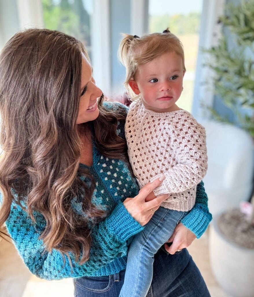 A woman with long brown hair holds a young child with light brown pigtails. Both are wearing cozy crocheted sweaters made from MJ's Off The Hook Designs, standing indoors near a window.
