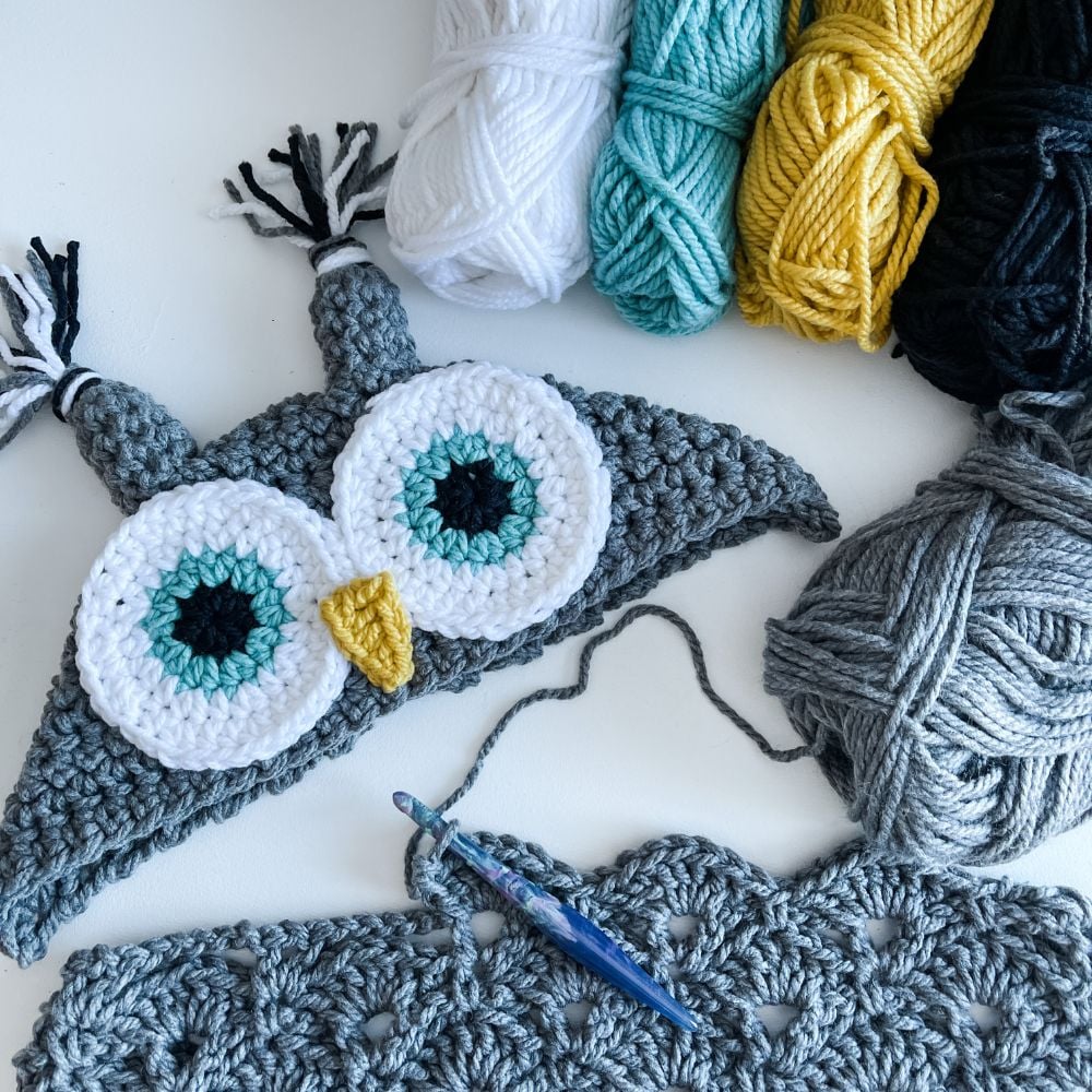 Crocheted owl face with yarn bundles in white, teal, yellow, black, and grey, plus a crochet hook and partially completed grey crochet piece.