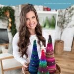 Woman holding a basket with three colorful crocheted Holiday Trees; festive decor and more crochet trees are visible in the background.