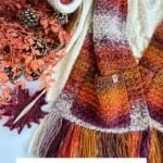 A crocheted autumn-colored pocket shawl with fringe is displayed on a cream sweater, surrounded by yarn, pinecones, leaves, and a maroon leaf-shaped dish. Text reads "Chunky Suzette Stitch Pocket Shawl." Perfect for cozy fall days.