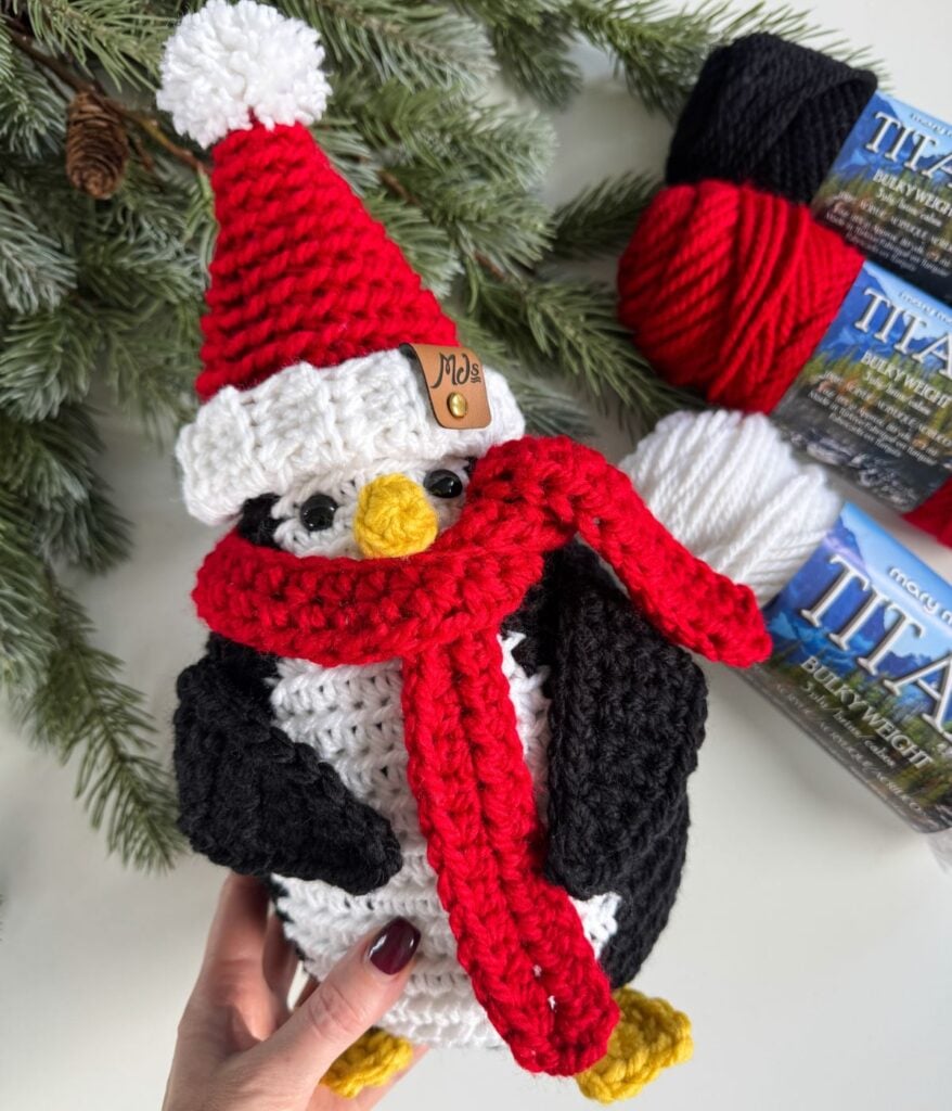 A hand holds a charming Santa Penguin, crocheted with a red scarf and Santa hat. Yarn balls and pine branches decorate the background.