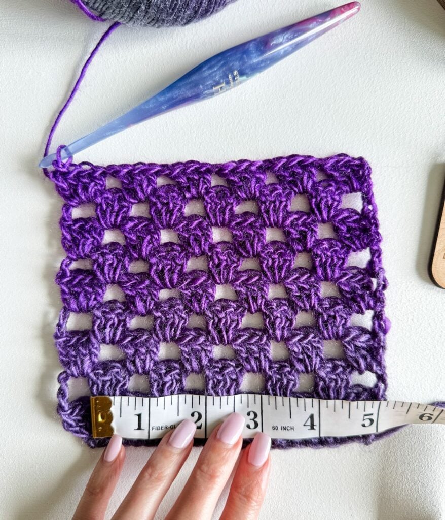 A purple crocheted square is measured with a tape measure, showing about 5 inches across—a great example for a beginners guide to crochet or checking your crochet gauge. A crochet hook and yarn are visible in the background.