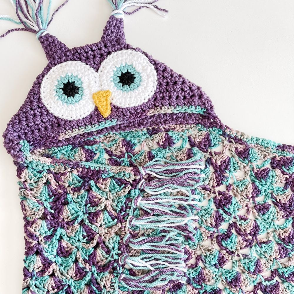 Crocheted blanket shaped like an owl, featuring purple, teal, and white yarn, with large eyes, a yellow beak, and fringe detail.