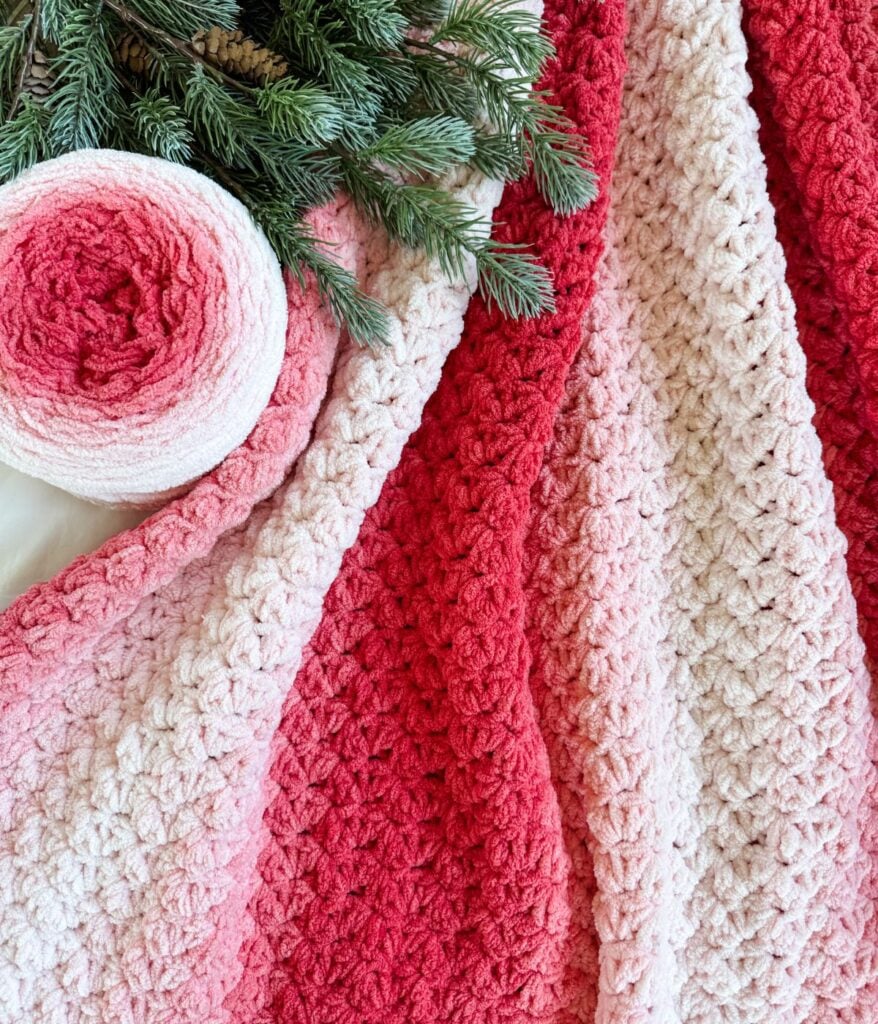 A soft, textured 2-day throw crochet blanket with red and pink ombre stripes is displayed next to a matching yarn ball and pine branches.