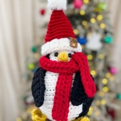 A Santa Penguin, crocheted with care and sporting a red Santa hat and matching scarf, is held in front of a blurred Christmas tree glowing with festive lights and ornaments.
