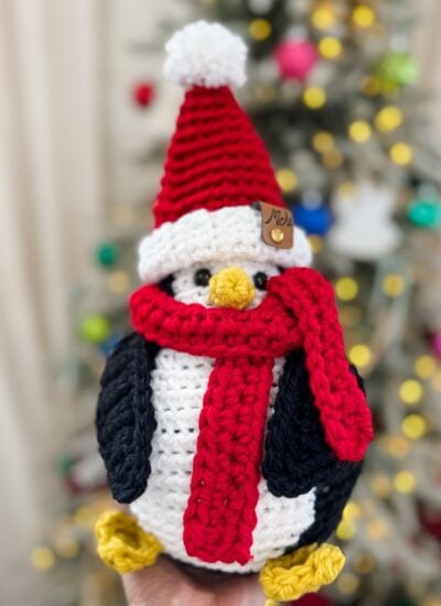A Santa Penguin, crocheted with care and sporting a red Santa hat and matching scarf, is held in front of a blurred Christmas tree glowing with festive lights and ornaments.