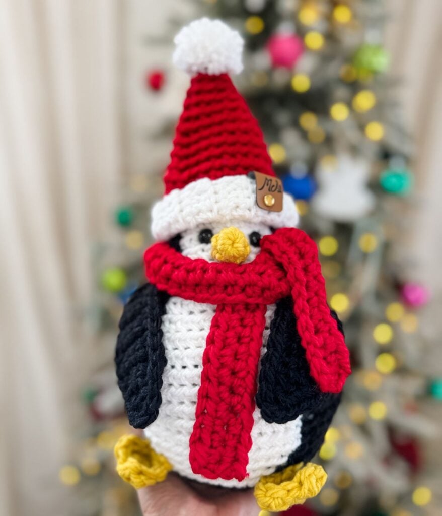 A Santa Penguin, crocheted with care and sporting a red Santa hat and matching scarf, is held in front of a blurred Christmas tree glowing with festive lights and ornaments.