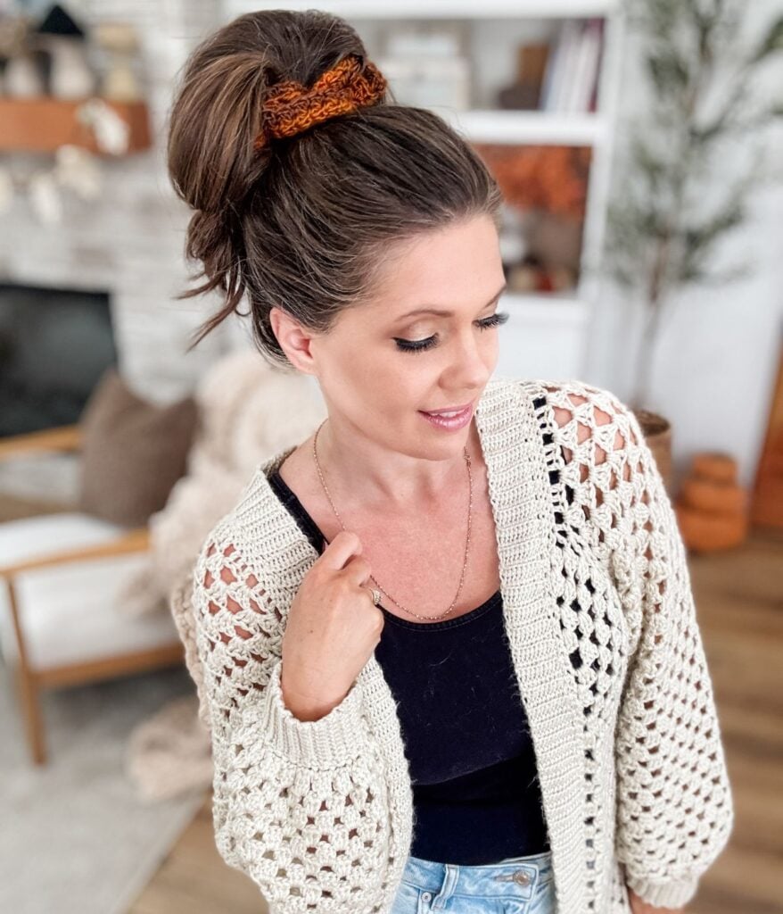 Woman with brunette hair in a high bun secured with a crochet scrunchie, wearing a cream cardigan over a black top, stands in a cozy, modern living room. The chic accessory features the Suzette Stitch for added texture and style.