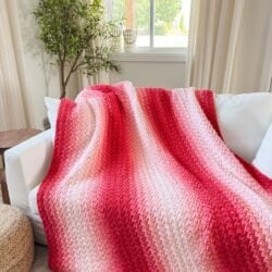 A red and pink gradient 2-day throw knit blanket draped over a white sofa in a bright, modern living room with a potted plant and window in the background.