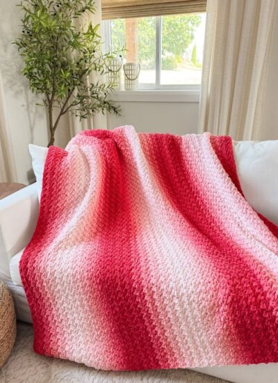 A red and pink gradient 2-day throw knit blanket draped over a white sofa in a bright, modern living room with a potted plant and window in the background.