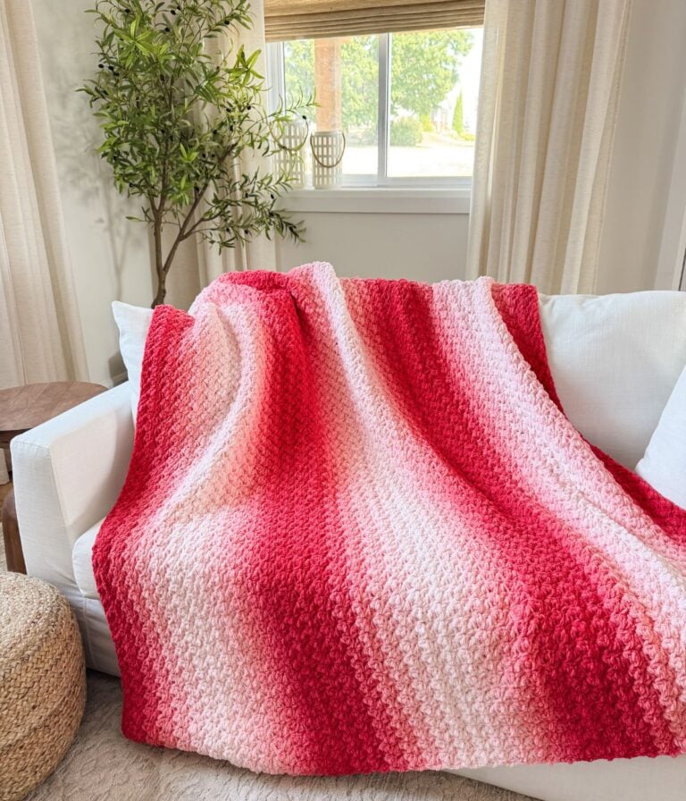 A red and pink gradient 2-day throw knit blanket draped over a white sofa in a bright, modern living room with a potted plant and window in the background.