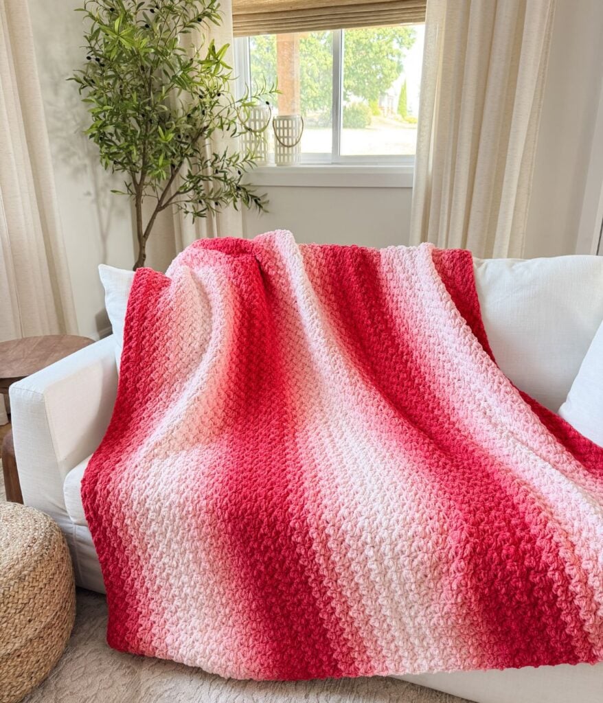 A red and pink gradient 2-day throw knit blanket draped over a white sofa in a bright, modern living room with a potted plant and window in the background.