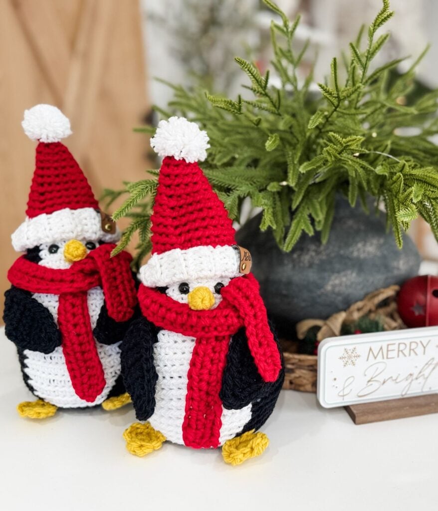 Two Santa Penguin crocheted figures, wearing red hats and scarves, are displayed next to a potted plant and a “Merry & Bright” sign.