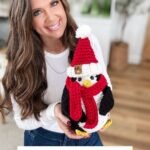 Woman holding a crocheted Santa Penguin wearing a festive hat and scarf, with text overlay offering a free Christmas penguin crochet pattern and video.