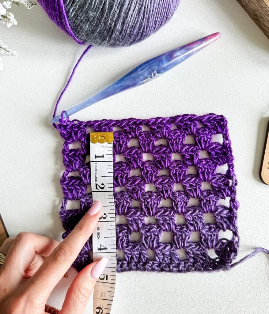 A hand holds a measuring tape against a purple crocheted square, with a crochet hook and yarn ball nearby—perfect for checking your crochet gauge as part of any beginner’s guide.