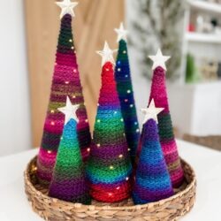 A woven basket holds six colorful, knitted cone-shaped trees with star toppers and tiny string lights, resembling the cozy charm of a pocket shawl.