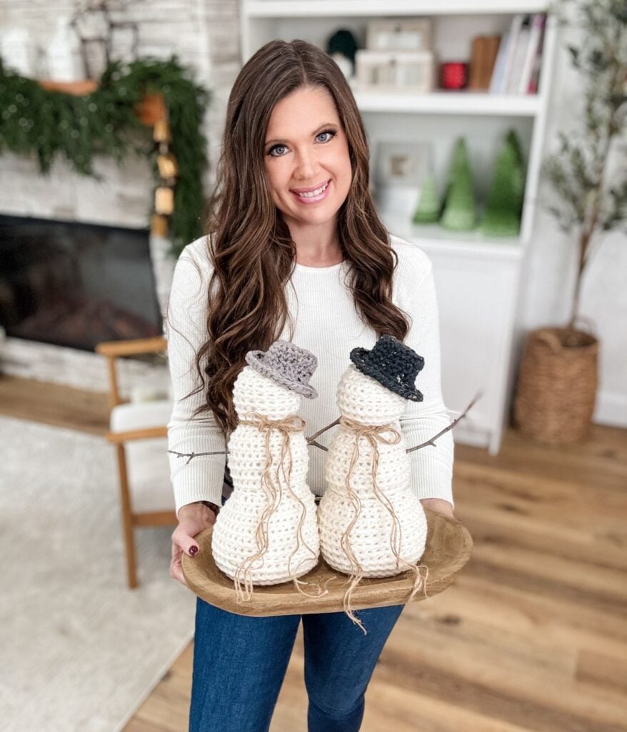 A woman in a white sweater holds a wooden tray with two crocheted snowmen from MJ's Off The Hook Designs in a cozy, decorated living room.